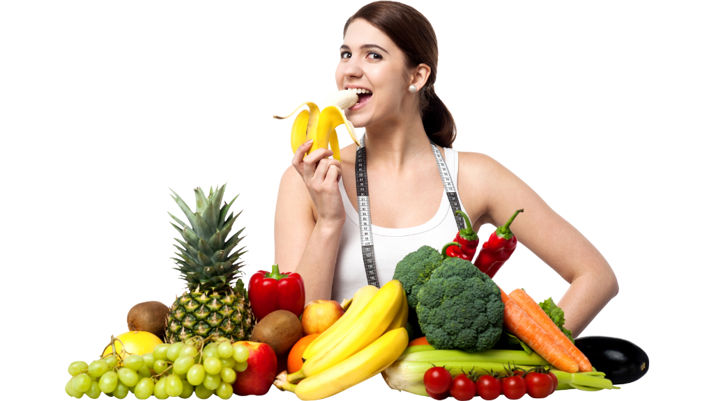 A woman eating healthily with a banana by a table full of fruit and veg